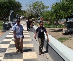 Picture of a woman in Timor Leste who is blind walking along a footpath at the waterfront with a mobility cane. A male orientation and mobility trainer is walking alongside her.